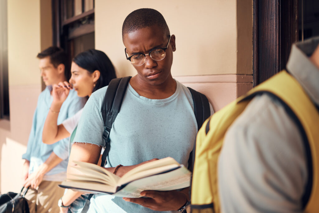 Student z Nigerii czyta książkę z plecakiem na plecach opierając się o ścianę